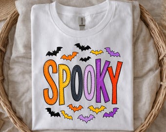 May include: A white short-sleeved t-shirt featuring the word SPOOKY in large, colorful, stitched-style letters. The letters are orange, yellow, dark gray, light purple, and red. Small black, orange, yellow, and purple bat silhouettes surround the text.