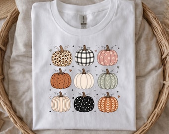May include: A white short-sleeved t-shirt featuring a graphic print of nine pumpkins arranged in a three-by-three grid. Each pumpkin displays a unique pattern, including leopard print, black and white plaid, polka dots, stripes, and floral motifs.