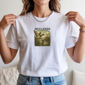 May include: A white short-sleeve t-shirt featuring a central graphic of two mallard ducks in a marshy environment with tall reeds and the text WETLANDS printed in black above the illustration. The design is framed within a rounded rectangle.