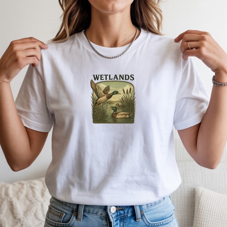 May include: A white short-sleeve t-shirt featuring a central graphic of two mallard ducks in a marshy environment with tall reeds and the text WETLANDS printed in black above the illustration. The design is framed within a rounded rectangle.