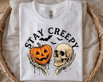 May include: A white t-shirt featuring a graphic print of two skeletal hands holding an orange jack-o'-lantern and a human skull. Above the graphic, the text "STAY CREEPY" is printed in black, accompanied by small black silhouettes of flying bats.