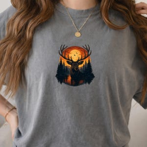 May include: A charcoal gray t-shirt featuring a central graphic of a deer with large antlers standing in a lake. The background shows a dark forest of pine trees silhouetted against a large, bright orange sunset with small birds flying in the sky.