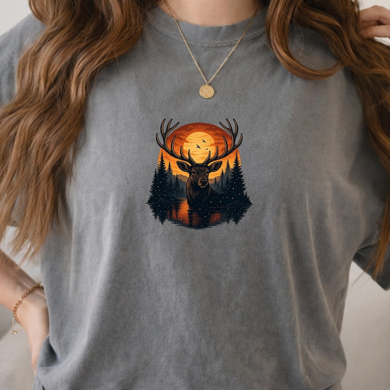 May include: A charcoal gray t-shirt featuring a central graphic of a deer with large antlers standing in a lake. The background shows a dark forest of pine trees silhouetted against a large, bright orange sunset with small birds flying in the sky.