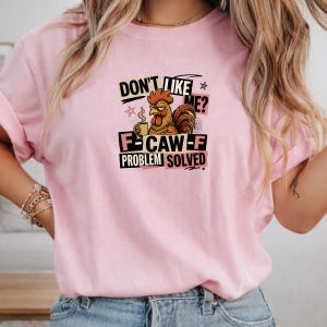 May include: A light pink t-shirt featuring a graphic of a brown rooster holding a coffee mug. The text reads: DON'T LIKE ME? F-CAW-F PROBLEM SOLVED. The design includes black and pink rectangular accents and small star symbols surrounding the rooster.
