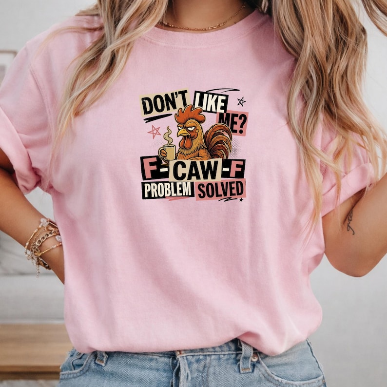 May include: A light pink t-shirt featuring a graphic of a brown rooster holding a coffee mug. The text reads: DON'T LIKE ME? F-CAW-F PROBLEM SOLVED. The design includes black and pink rectangular accents and small star symbols surrounding the rooster.