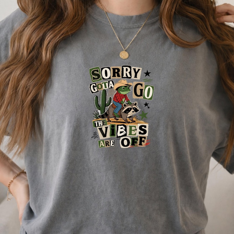 May include: A charcoal gray t-shirt featuring a graphic print of a frog wearing a cowboy hat and red plaid shirt riding a raccoon. The design includes a green cactus, small stars, and the text: SORRY GOTA GO THE VIBES ARE OFF in block letters.