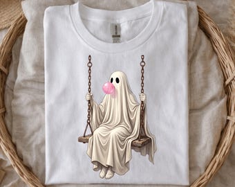 May include: A white t-shirt featuring a graphic illustration of a ghost draped in a white sheet, sitting on a wooden swing suspended by metal chains. The ghost is blowing a round, bright pink bubble with bubblegum in front of its face.