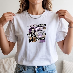 May include: A white t-shirt featuring a graphic of a skeleton with purple hair, sunglasses, and a leopard print top, holding a drink and making a peace sign. The text reads: IF MY personalities OFFEND YOU, I'M SURE ONE OF them CAN TELL YOU WHERE TO GO.