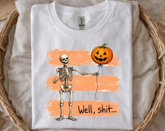 May include: A white t-shirt featuring a graphic of a skeleton holding a string attached to an orange jack-o'-lantern pumpkin balloon. Behind the skeleton are three horizontal orange brush strokes. The text "Well, shit..." is printed in black.