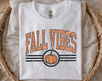 May include: A white short-sleeve t-shirt featuring the text FALL VIBES in orange block letters with a distressed finish. Below the text is a graphic of an orange pumpkin centered within a dark gray circle, flanked by three horizontal lines.