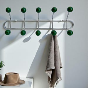 May include: A chrome wall-mounted coat rack with eight green ball-shaped hooks. A beige towel hangs from one of the hooks. The rack is mounted on a white wall. A brown hat sits on a white surface below the rack.