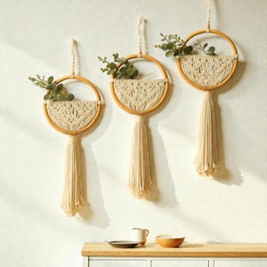 May include: Three macrame wall hangings with wooden hoops and cream-colored tassels. Each features a woven design and holds faux greenery. The hangings are suspended against a white wall, creating a bohemian aesthetic.
