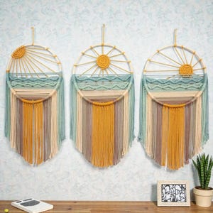 Könnte beinhalten: Drei handgefertigte Wandbehänge mit Sonnen- und Ozean-Design. Jedes Stück hat einen kreisförmigen Rahmen mit einer gelben Sonne, blauen und beigen Wellen und vertikalen Strängen in Gelb-, Beige- und Blautönen. Wanddekoration.