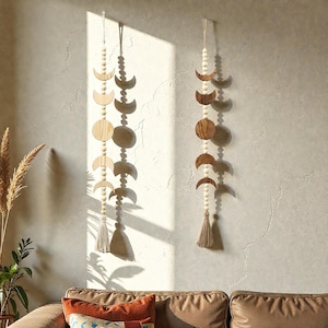 May include: Two wooden moon phase wall hangings. Each features a string of wooden beads, crescent moons, and circular shapes in varying shades of brown and beige. A beige tassel hangs at the bottom of each piece. The wall hangings are suspended against a neutral-toned wall.