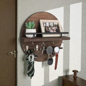 May include: A circular, dark wood wall shelf with a small shelf, a black metal bar, and hooks. It displays a small potted plant, a framed photo, keys, sunglasses, a watch, and a decorative tie. The shelf is mounted on a light grey wall.