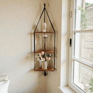 Puede incluir: Estantería esquinera colgante con tres niveles de madera, suspendida por una cuerda negra. Las estanterías exhiben un busto blanco, una botella de vidrio transparente y un jarrón blanco, cada uno con flores. La estantería está cerca de una ventana.