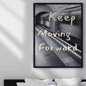 Peut inclure: Impression en noir et blanc encadrée d'un train de métro flou dans un tunnel. L'inscription dorée "Keep Moving Forward" est superposée. L'image est affichée au-dessus d'un lit avec des oreillers blancs.