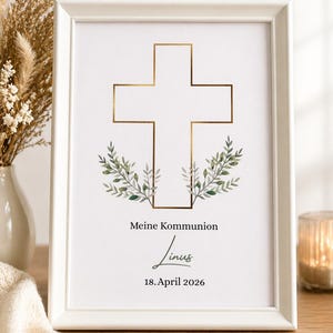 May include: A framed print with a gold cross design, green leaf accents, and the text "Meine Kommunion Linus 18. April 2026." The white frame surrounds the print, which is displayed on a wooden surface.