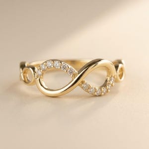 Pode incluir: Um anel de ouro com um design de símbolo de infinito. A banda é feita de ouro amarelo polido, com um lado do laço do infinito adornado com uma série de pequenas pedras transparentes de corte redondo ao longo da curva superior.