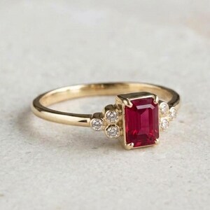 May include: A gold ring featuring a central emerald-cut red ruby gemstone held in a prong setting, flanked by three small round white diamonds on each side of the band, creating a symmetrical design with a polished yellow gold metal finish.