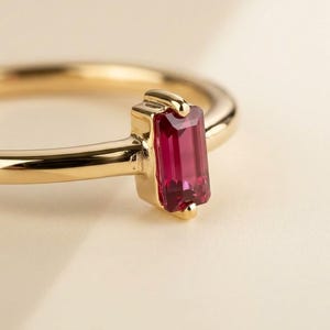 Op de afbeelding: Een gouden ring met een enkele, smaragdgeslepen dieproze edelsteen die verticaal is gezet in een gepolijste gouden zetting met pootjes. De ringband is glad en rond en toont een strak ontwerp dat de levendige kleur van de steen benadrukt.