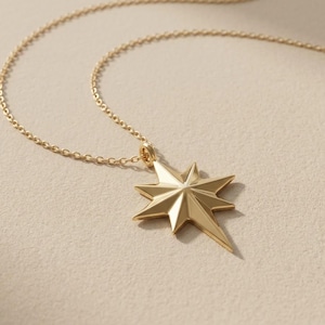 May include: A gold-toned necklace featuring a polished eight-pointed star pendant suspended from a delicate matching chain. The star charm has faceted surfaces that create a dimensional look, with one elongated point extending downward from the centre.