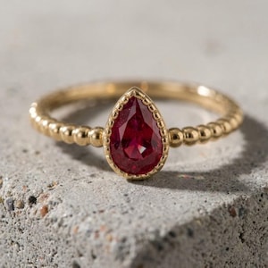 Pode incluir: Um anel de ouro com uma pedra preciosa vermelha em forma de pera engastada num bisel de contas. A banda do anel é composta por uma série de pequenas esferas de ouro ligadas entre si, criando um aspeto texturado e uniforme.