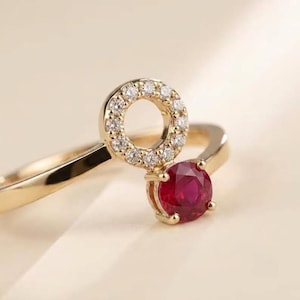 May include: A gold ring featuring a circular halo of small white diamonds and a single round-cut deep red ruby stone. The band is smooth and polished, connecting to the circular setting that holds the diamonds and the prominent red gemstone.