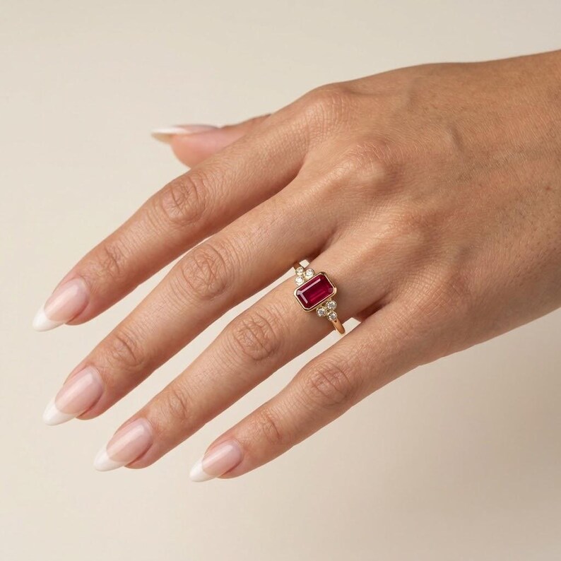 Peut inclure: Une bague en or orn&eacute;e d'une pierre rouge rectangulaire taille &eacute;meraude mont&eacute;e en serti clos. La pierre centrale est entour&eacute;e de trois petites pierres blanches rondes de chaque c&ocirc;t&eacute;, dispos&eacute;es en grappe sur le fin anneau en or poli.