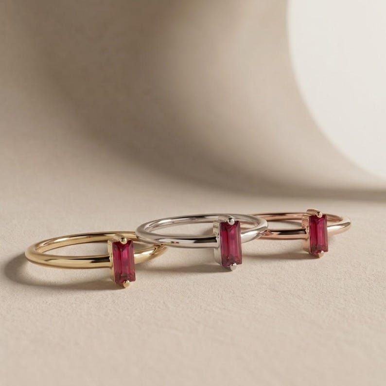 Puede incluir: Tres anillos finos de metal dispuestos en fila, cada uno con una piedra preciosa rectangular de color rojo intenso en talla baguette montada verticalmente. Los anillos son de oro amarillo, plata y oro rosa, con un dise&ntilde;o de banda minimalista.