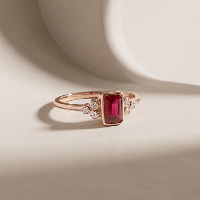 Peut inclure: Une bague en or rose orn&eacute;e d'une pierre rouge centrale taille &eacute;meraude sertie clos, entour&eacute;e de trois petits diamants ronds blancs de chaque c&ocirc;t&eacute;. L'anneau est lisse et poli, pr&eacute;sentant un design &eacute;pur&eacute; avec une pierre rouge &eacute;clatante.