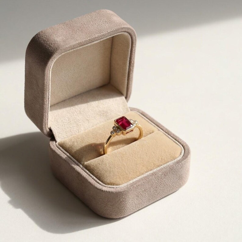 Peut inclure: Une bague en or orn&eacute;e d'une pierre pr&eacute;cieuse rouge rectangulaire au centre, flanqu&eacute;e de trois petites pierres claires rondes de chaque c&ocirc;t&eacute;. La bague est pr&eacute;sent&eacute;e dans un &eacute;crin &agrave; bijoux ouvert, doubl&eacute; de velours beige, avec un ext&eacute;rieur beige.
