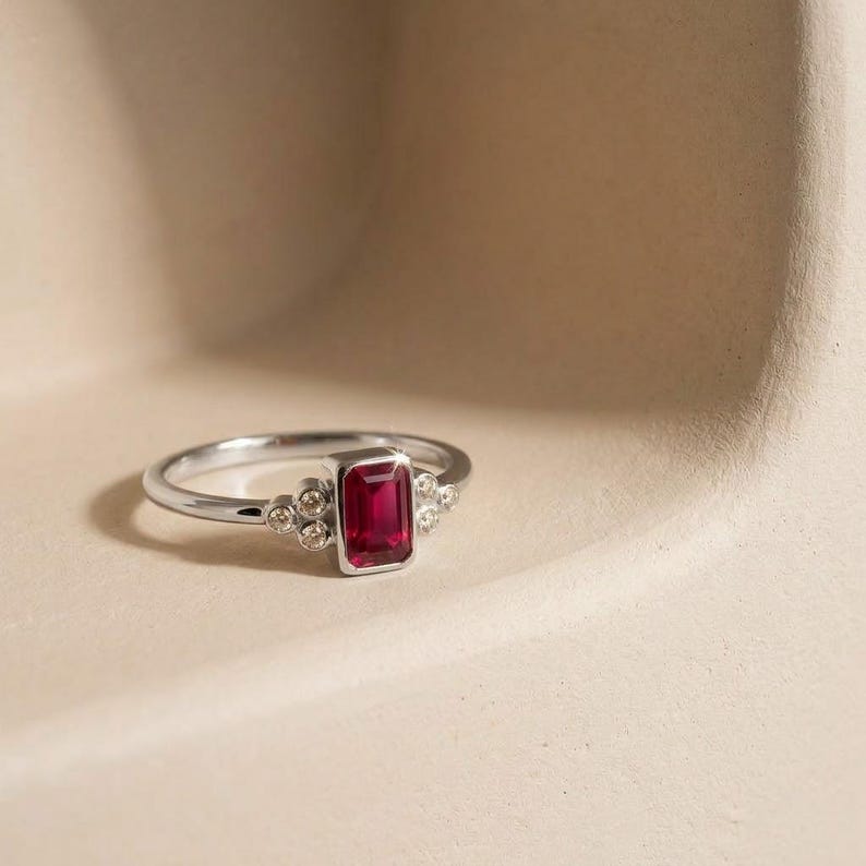 Peut inclure: Une bague en argent orn&eacute;e d'une pierre rouge centrale taille &eacute;meraude sertie clos. L'anneau est agr&eacute;ment&eacute; de chaque c&ocirc;t&eacute; par un groupe de trois petites pierres rondes et transparentes, offrant un design &eacute;quilibr&eacute; au fini m&eacute;tallique poli.