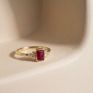 Peut inclure: Une bague en or avec un rubis rouge central de taille &eacute;meraude rectangulaire dans un serti clos, flanqu&eacute; de trois petits diamants blancs ronds de chaque c&ocirc;t&eacute;, sur un anneau en or poli qui s'affine vers l'arri&egrave;re de la bague.