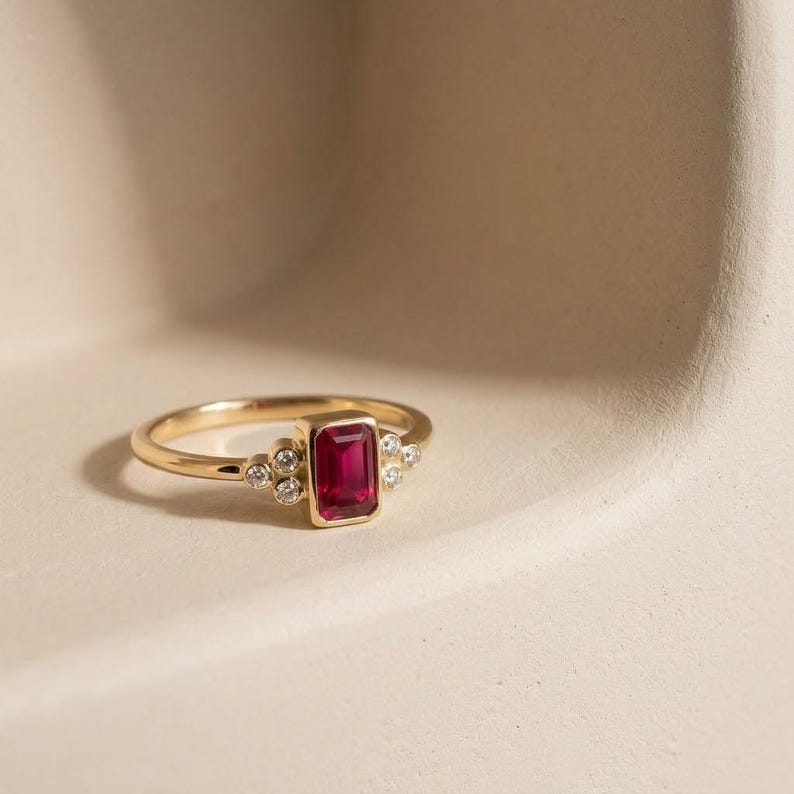 Peut inclure: Une bague en or avec un rubis rouge central de taille &eacute;meraude rectangulaire dans un serti clos, flanqu&eacute; de trois petits diamants blancs ronds de chaque c&ocirc;t&eacute;, sur un anneau en or poli qui s'affine vers l'arri&egrave;re de la bague.