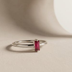 Puede incluir: Un anillo de tono plateado que presenta una &uacute;nica piedra preciosa rectangular de corte baguette, orientada verticalmente, en un color rosa intenso y vibrante. La banda es lisa, pulida y delgada, sosteniendo la piedra en un engaste sencillo de cuatro garras.