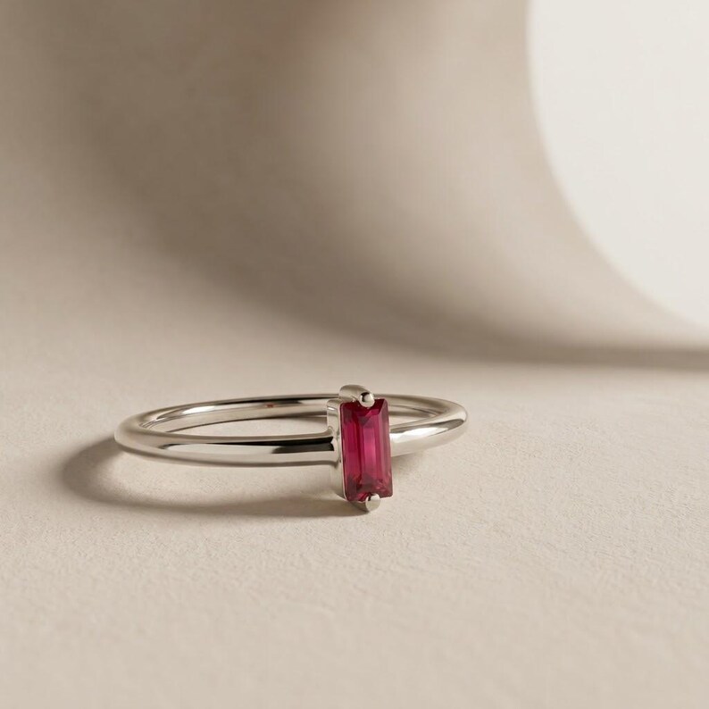 Puede incluir: Un anillo de tono plateado que presenta una &uacute;nica piedra preciosa rectangular de corte baguette, orientada verticalmente, en un color rosa intenso y vibrante. La banda es lisa, pulida y delgada, sosteniendo la piedra en un engaste sencillo de cuatro garras.