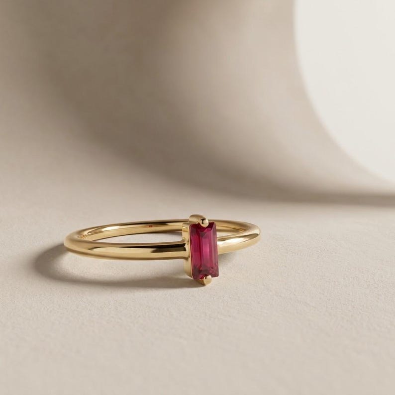 Puede incluir: Un anillo de oro con una &uacute;nica piedra preciosa roja de corte baguette rectangular orientada verticalmente, engastada en una banda de oro pulido sencilla. El anillo tiene un cuerpo redondeado y liso con un engaste de cuatro garras.