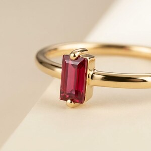 Puede incluir: Un anillo de oro con una sola piedra preciosa roja de corte baguette rectangular, engastada verticalmente en un engaste de bisel de oro pulido. La banda es fina y lisa, hecha de oro amarillo, destacando la piedra roja vibrante.