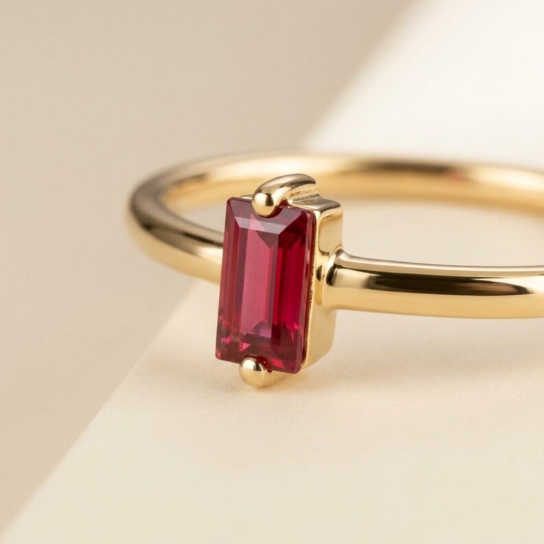 Puede incluir: Un anillo de oro con una sola piedra preciosa roja de corte baguette rectangular, engastada verticalmente en un engaste de bisel de oro pulido. La banda es fina y lisa, hecha de oro amarillo, destacando la piedra roja vibrante.