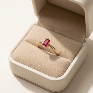Puede incluir: Un anillo de oro con una piedra preciosa roja de corte baguette rectangular engastada en una banda de metal sencilla. El anillo se presenta dentro de un joyero cuadrado de color crema con un forro interior de terciopelo beige suave.