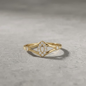 May include: A gold ring featuring a central marquise-shaped element encrusted with small round white stones. The band splits into two arms that meet at the center, with a single small round white stone set at each side where the arms converge.
