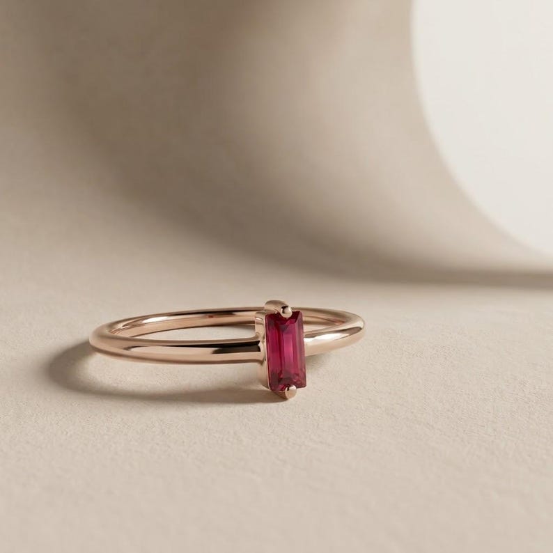 Puede incluir: Un anillo de oro rosa con una piedra preciosa de color rosa intenso, corte baguette rectangular y orientaci&oacute;n vertical, montada en una banda de metal pulido. El anillo presenta un dise&ntilde;o sencillo con un cuerpo liso y redondeado.