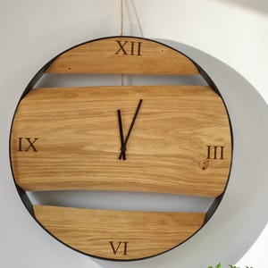 以下が含まれることがあります： 3時、6時、9時、12時の位置にローマ数字が配置された丸い木製の壁掛け時計。文字盤は明るい色の木材で、暗い金属フレームが付いています。時計の針は黒色です。白い壁に掛けられています。