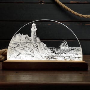 Pode incluir: Luz decorativa com uma gravura detalhada de um farol numa falésia, com um veleiro no mar. A luz está assente numa base de madeira e tem um topo curvo. O design evoca um tema náutico, com uma cena costeira.