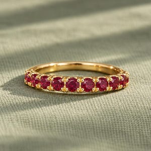 Könnte beinhalten: Ein goldener Ring mit einer durchgehenden Reihe runder, leuchtend roter Edelsteine, die in einer gemeinsamen Krappenfassung gefasst sind. Das Metall hat eine polierte Oberfläche und die Steine sind in Größe und Form einheitlich.