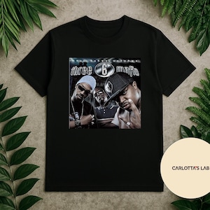 Peut inclure: T-shirt noir avec un graphisme du groupe de rap Three 6 Mafia. Le graphisme comprend le nom du groupe et la phrase "Most Known Unknown". L'image montre trois hommes portant des lunettes de soleil et des chapeaux.