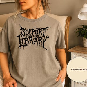 May include: A grey t-shirt featuring the black text "Support Your Local Library" in a gothic font. The shirt has a relaxed fit and is a neutral colour. The text is the main design element.
