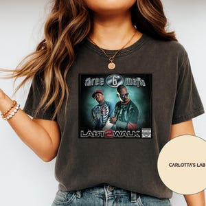 Peut inclure: T-shirt gris foncé avec la pochette de l'album "Last 2 Walk" de Three 6 Mafia. Le motif comprend deux hommes, le titre de l'album et le nom du groupe. Le t-shirt a un col rond et des manches courtes.