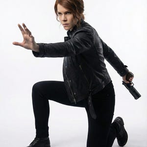 May include: A person kneels, wearing black trousers, boots, and a leather jacket, holding a handgun. The person has auburn hair and is positioned with one arm outstretched, as if in action, against a white backdrop.
